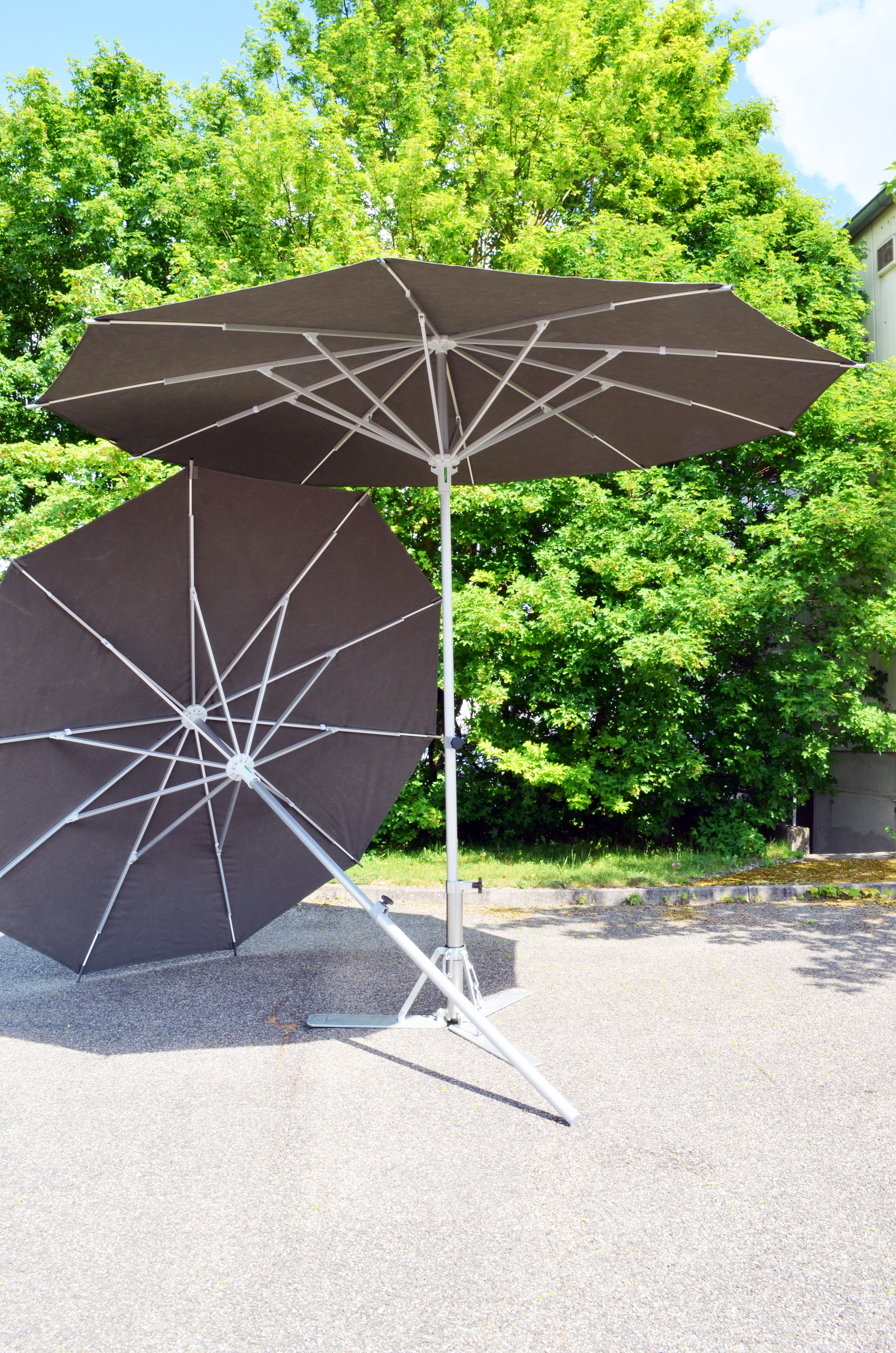 Zwei große schwarze Sonnenschirme mit weißem Metallgestell auf Asphalt vor grünen Bäumen