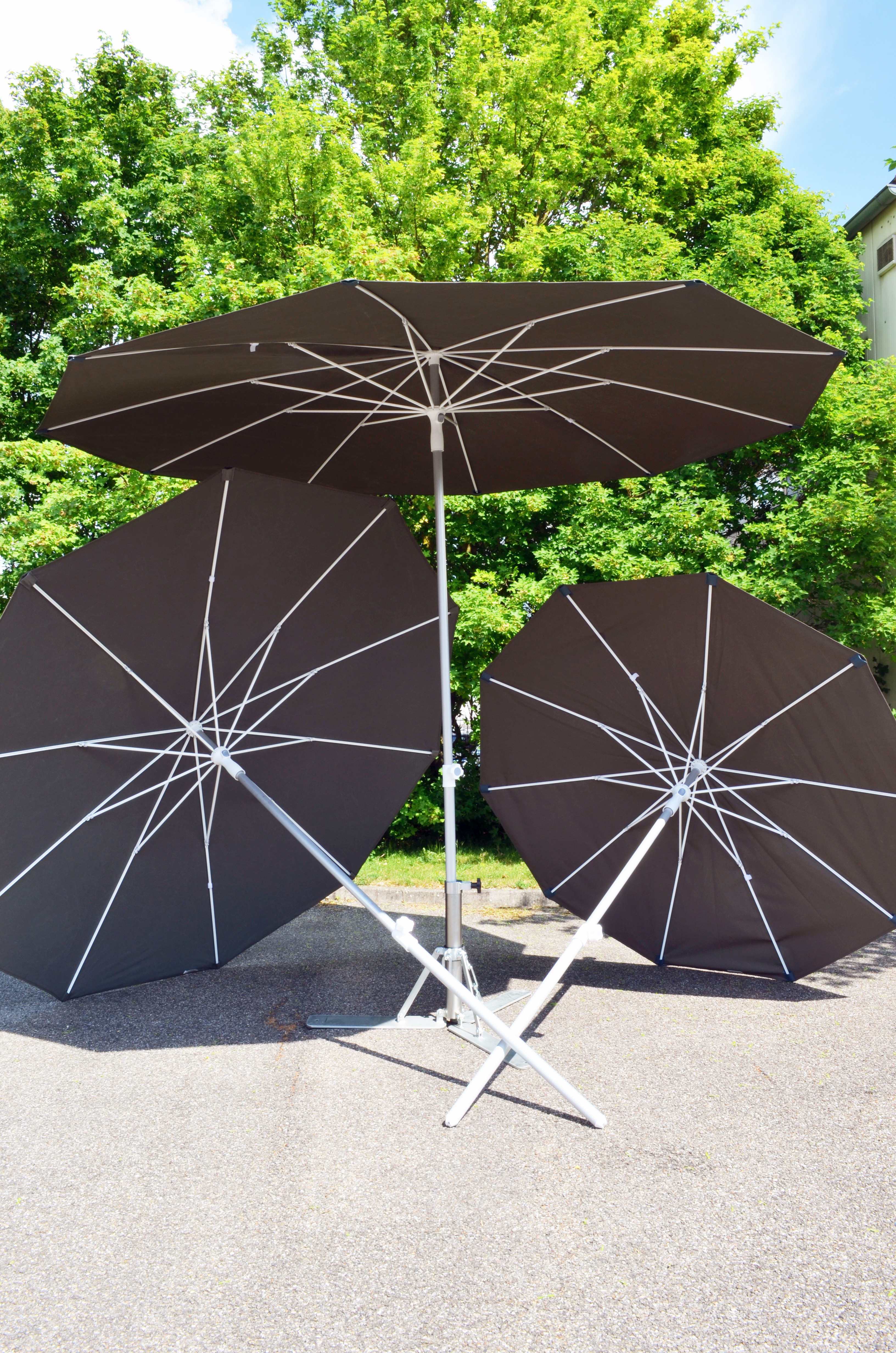 Drei schwarze Sonnenschirme mit weißem Gestell auf Asphalt vor grünen Bäumen