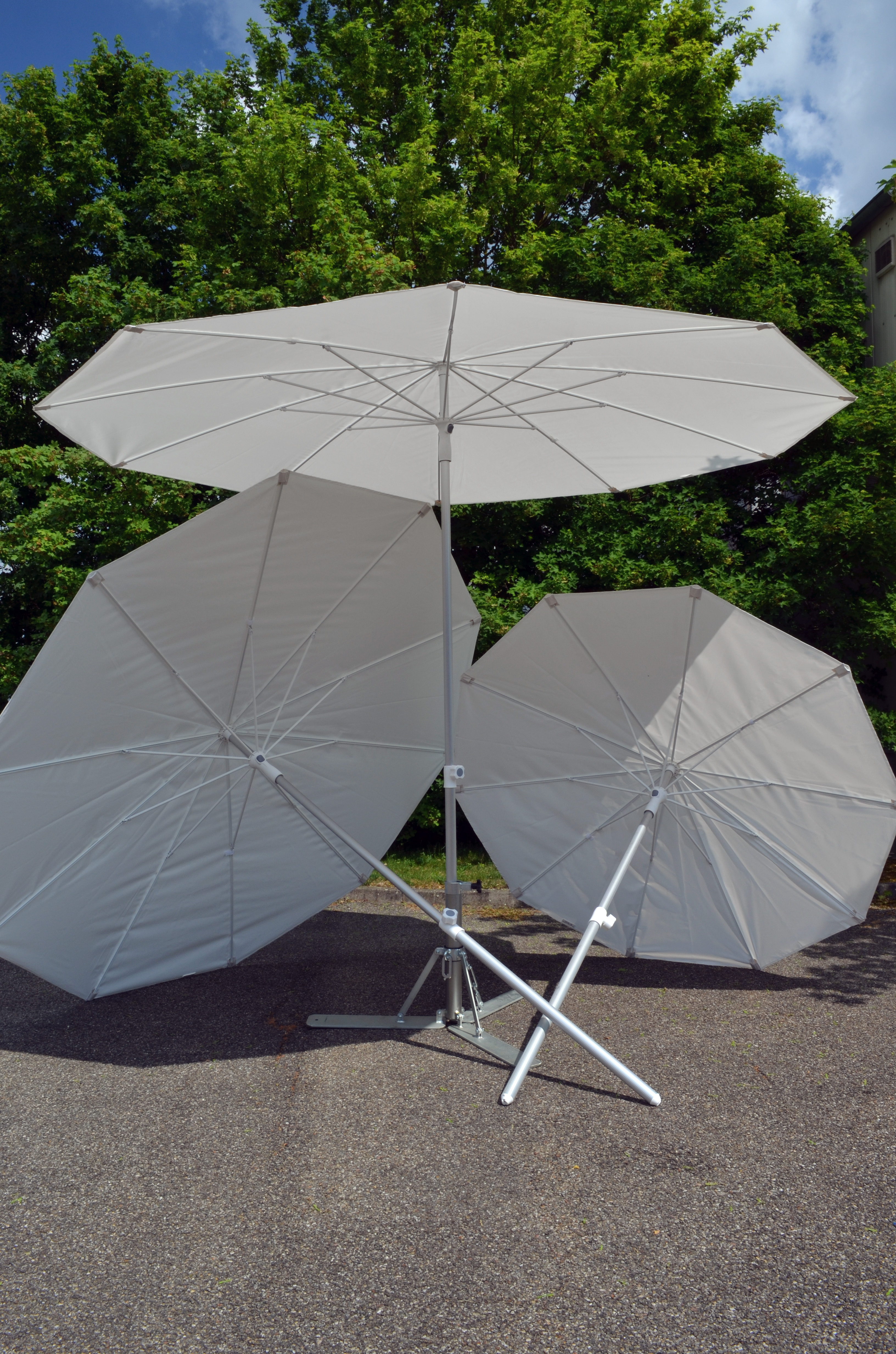 Drei weiße Sonnenschirme mit Metallgestell auf Asphalt vor grünen Bäumen
