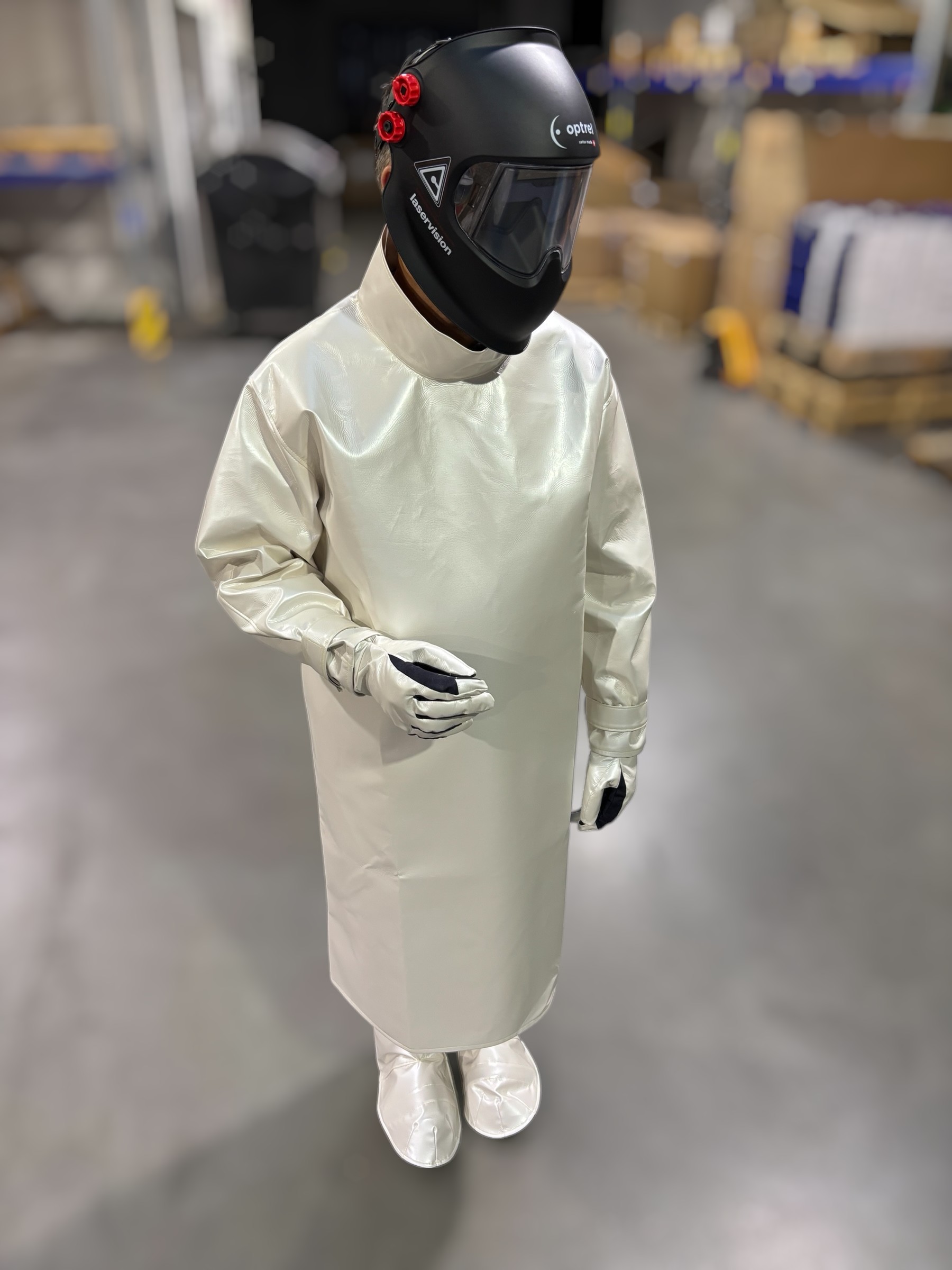Person steht in Schutzkleidung mit langem Kittel, Handschuhen, Überschuhen und einem schwarzen Laserhelm mit Sichtfenster und roten Bedienelementen