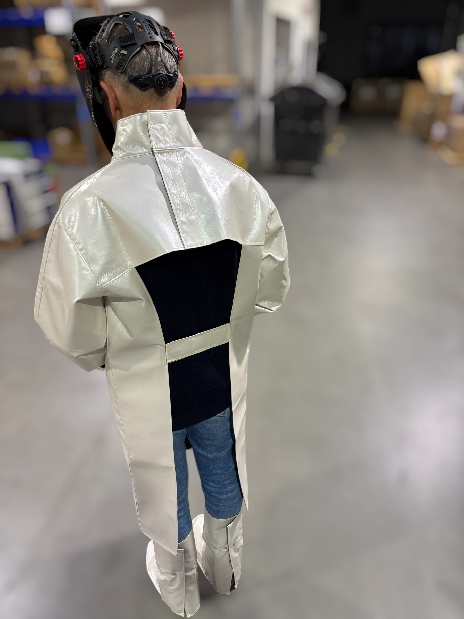 Person steht in Schutzkleidung mit langem Kittel, Handschuhen, Überschuhen und einem schwarzen Laserhelm mit Sichtfenster und roten Bedienelementen