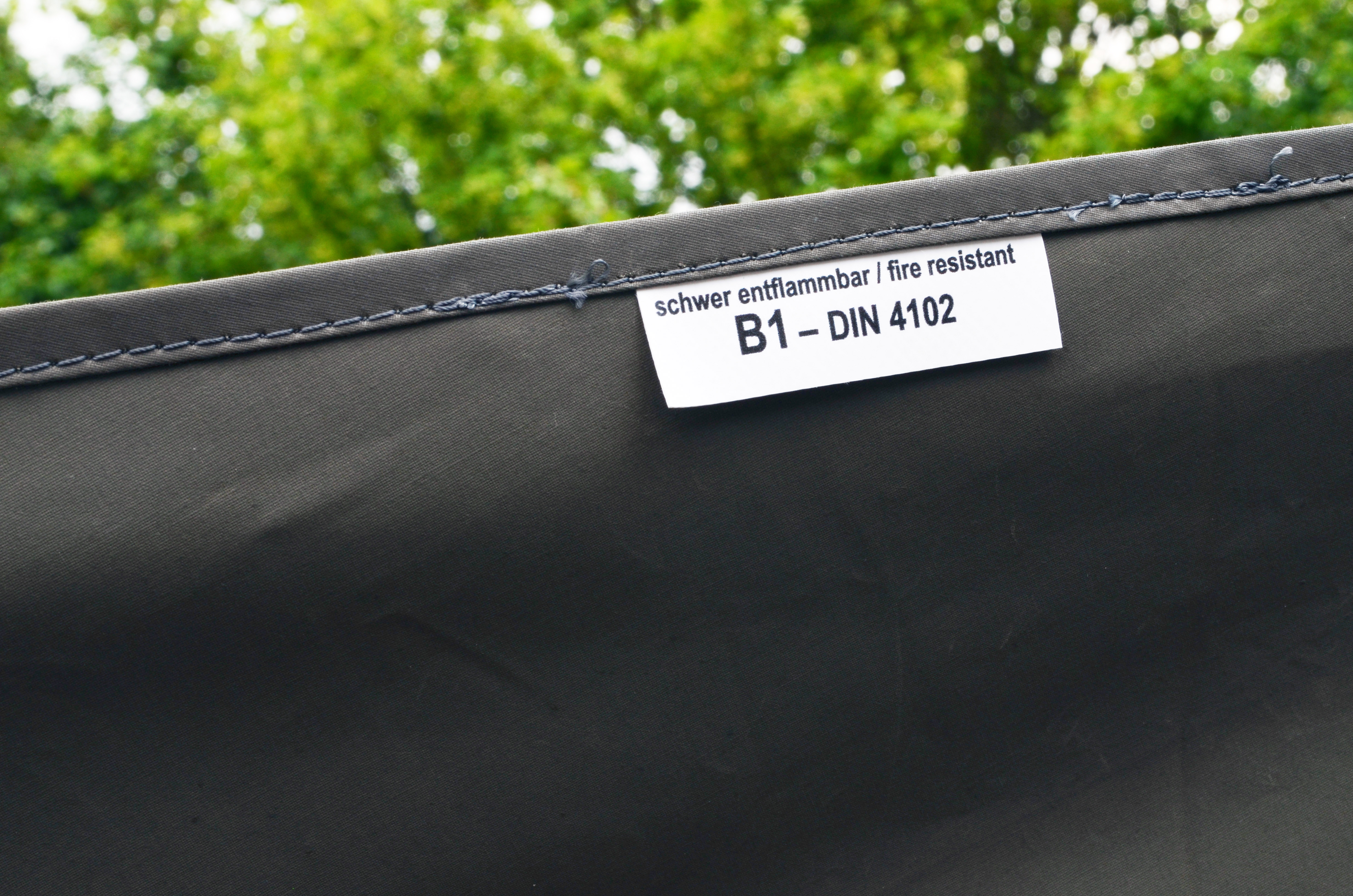 Schwarzer Stoff mit weißem Etikett 'schwer entflammbar | fire resistant B1 – DIN 4102' und sichtbarer Naht